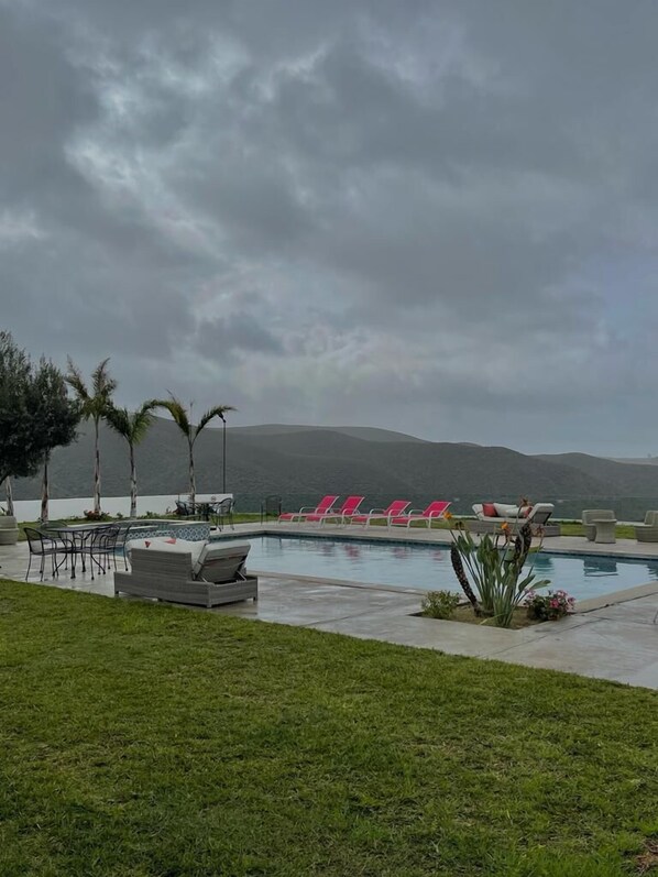 A pool area with lounge chairs and a table, surrounded by grass and palm trees, with a view of hills in the background. - Viñas del Castillo Hotel Boutique (Ensenada)