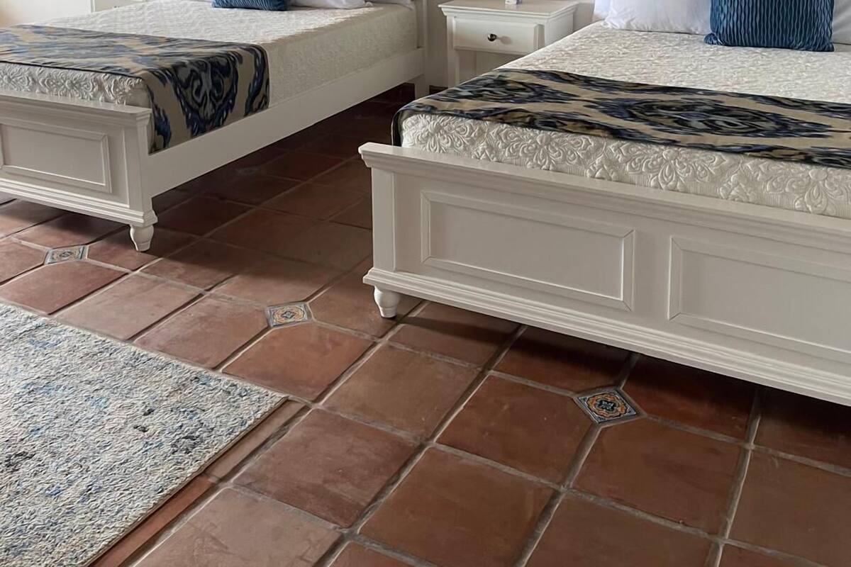Deux lits blancs avec des couvre-lits à motifs, une petite table de chevet avec une plante, et un sol carrelé.
