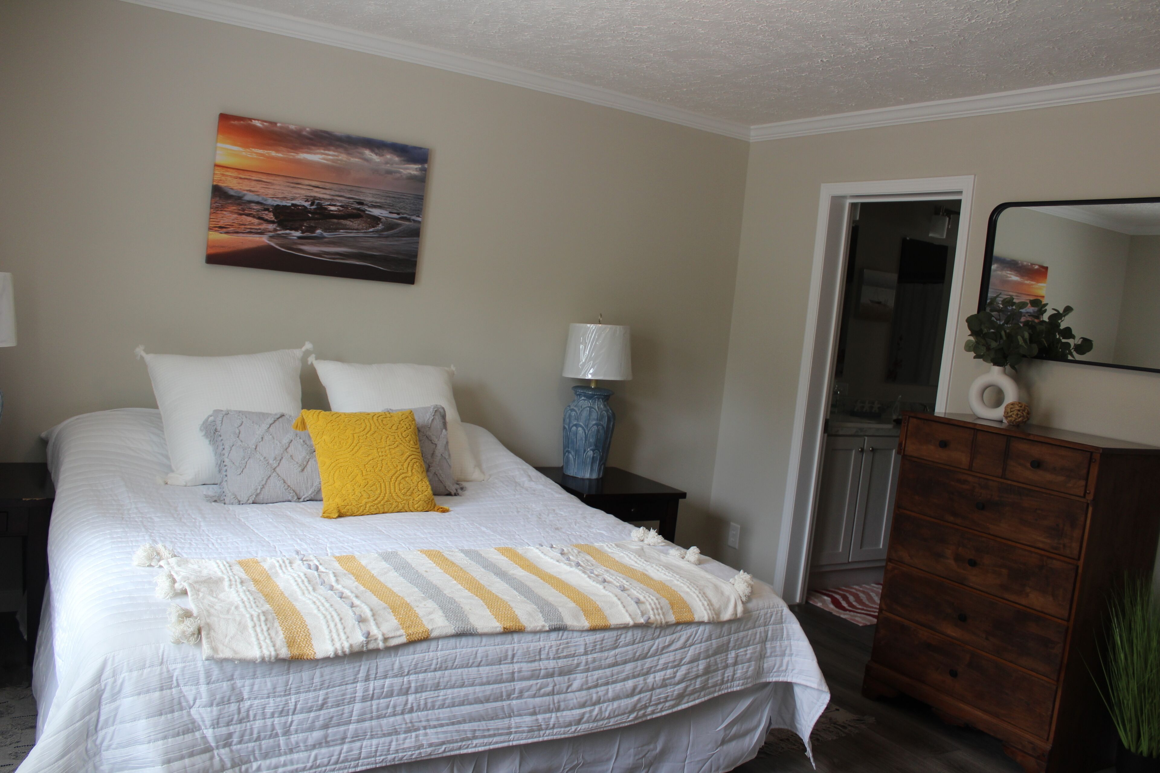 Une chambre à coucher avec un lit, une table de chevet, une commode, un miroir et un tableau accroché au mur.