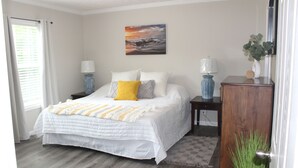 A bedroom with a bed, two bedside tables, a nightstand, a painting on the wall, a door, and a window with curtains.