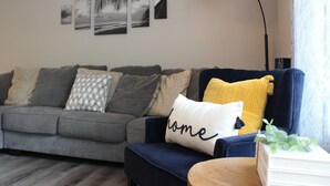 A living room with a grey sofa, a blue armchair, and a yellow pillow. There are wall art pieces above the sofa and a floor lamp to the right.