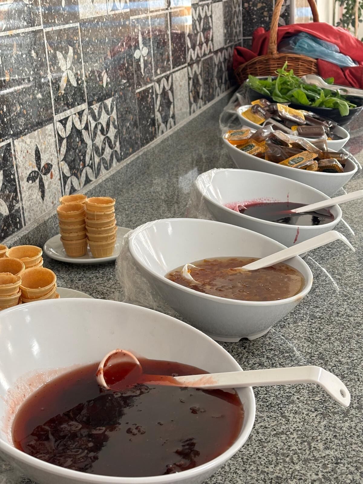 A buffet table with various food items and bowls, some covered with plastic.