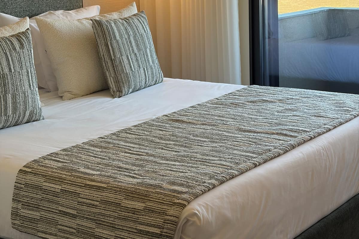 Ein Hotelzimmer mit einem großen Bett, einem Kopfteil, einem Nachttisch, einer Lampe und Blick auf eine Landschaft durch das Fenster.