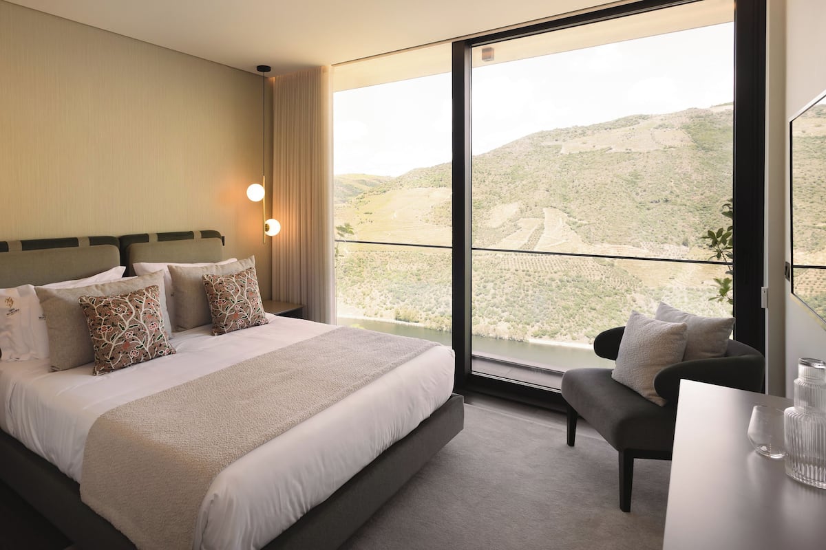 Ein Hotelzimmer mit einem großen Bett, zwei Nachttischlampen, Blick auf eine hügelige Landschaft, einem kleinen Schreibtisch und einem Sessel.