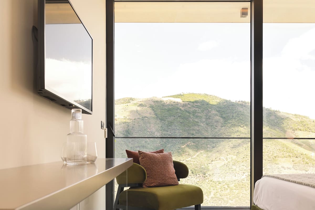 Ein modernes Schlafzimmer mit großem Fenster, einem Bett, einem grünen Sessel, einem kleinen Tisch und einem an der Wand befestigten Fernseher.