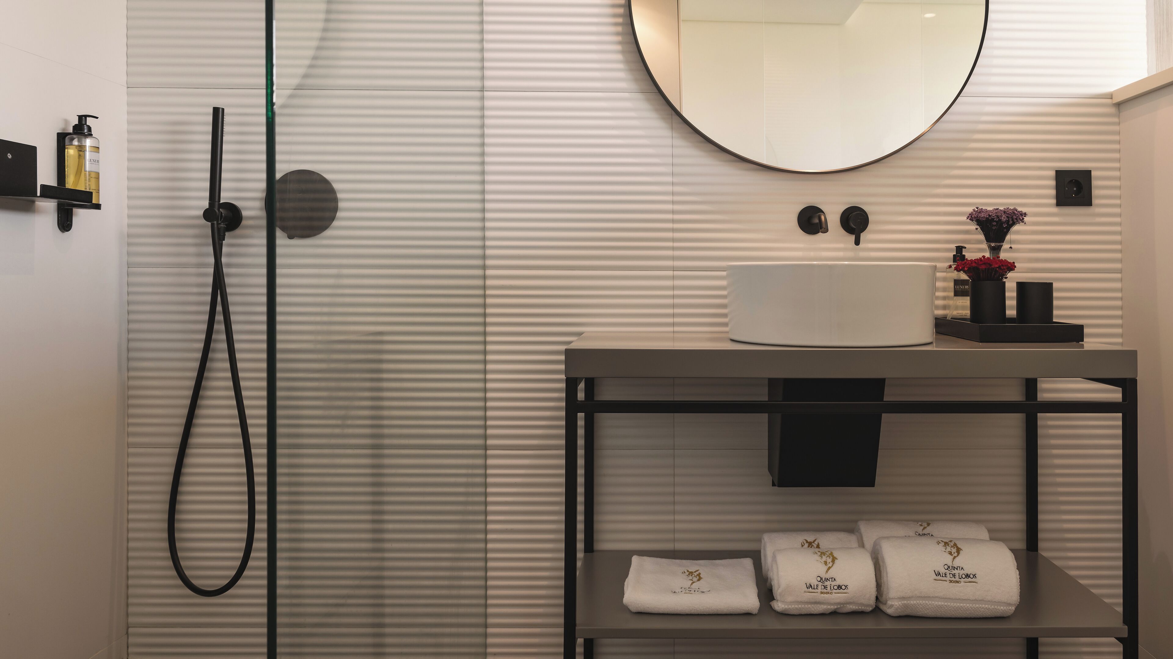 A modern bathroom with a shower, a round mirror, a sink with a white basin, and a shelf with towels.