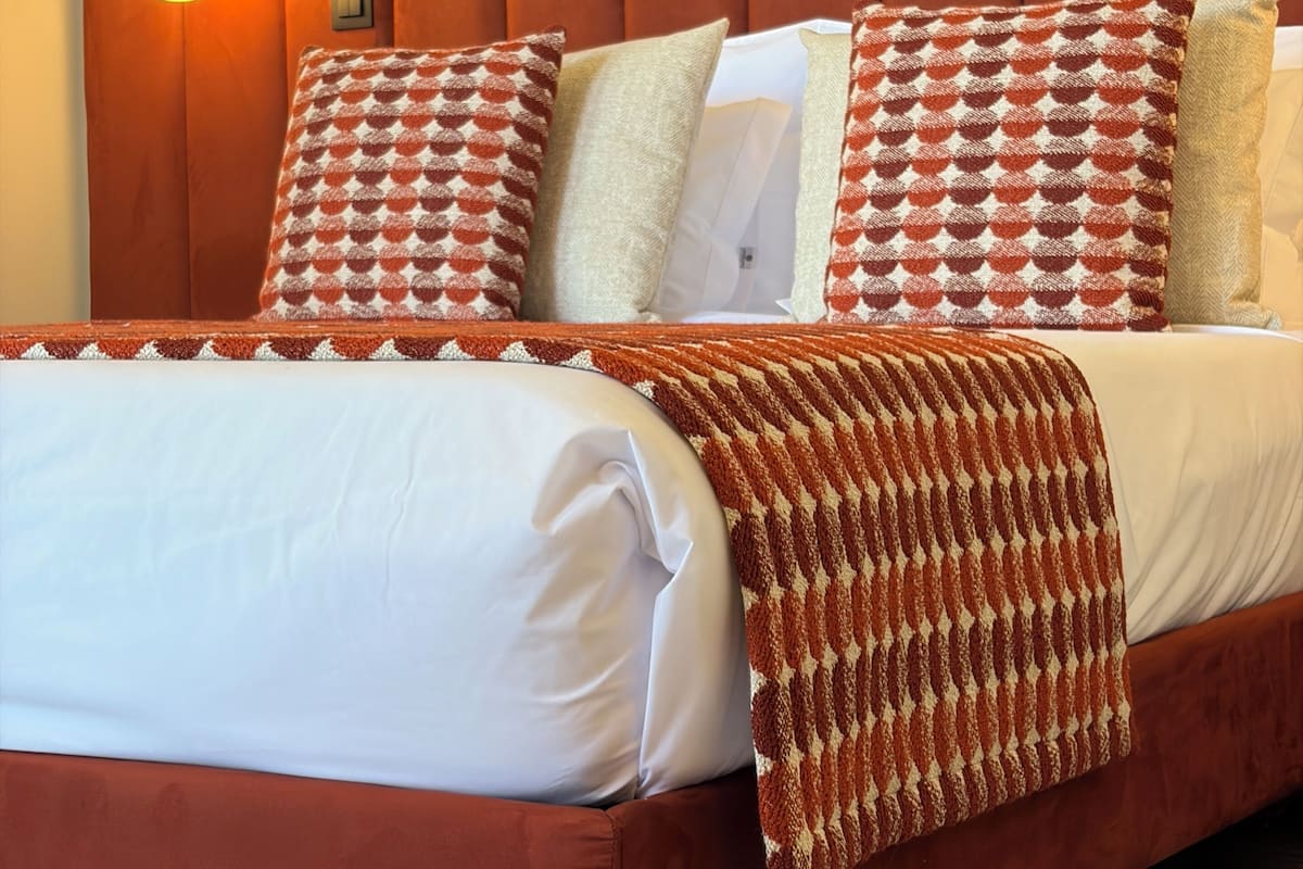 Ein Bett mit Holz-Kopfstück, weißer Bettwäsche, gemusterten Kissen und einer braunen Tagesdecke.