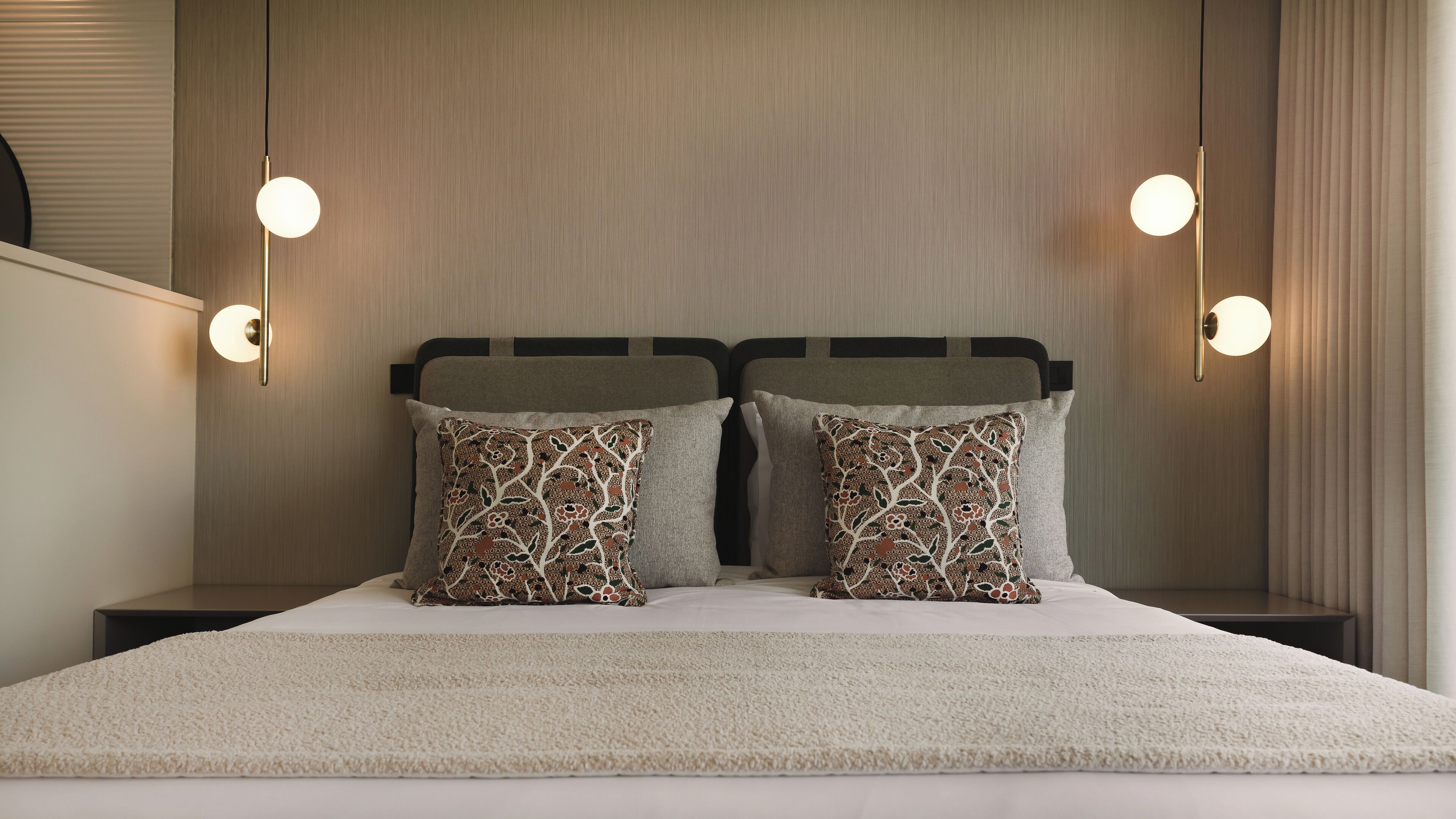 A neatly made bed with a beige blanket, two patterned pillows, and a headboard with a light wood finish. There are two bedside lamps with round shades on either side of the bed.