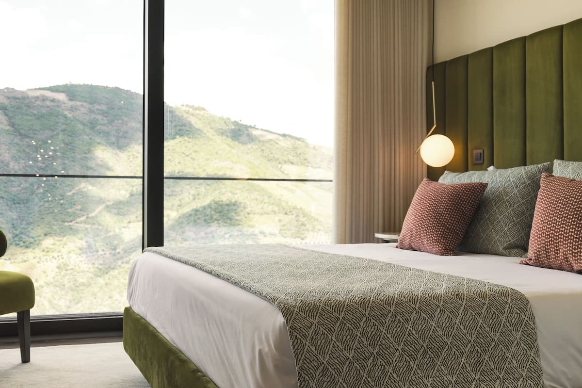 Ein Hotelzimmer mit großem Fenster und Blick auf eine gebirgige Landschaft. Es beinhaltet ein Bett mit gemusterten Betttextilien, einen mit Stoff bezogenen grünen Sessel und einen Nachttisch mit Lampe.