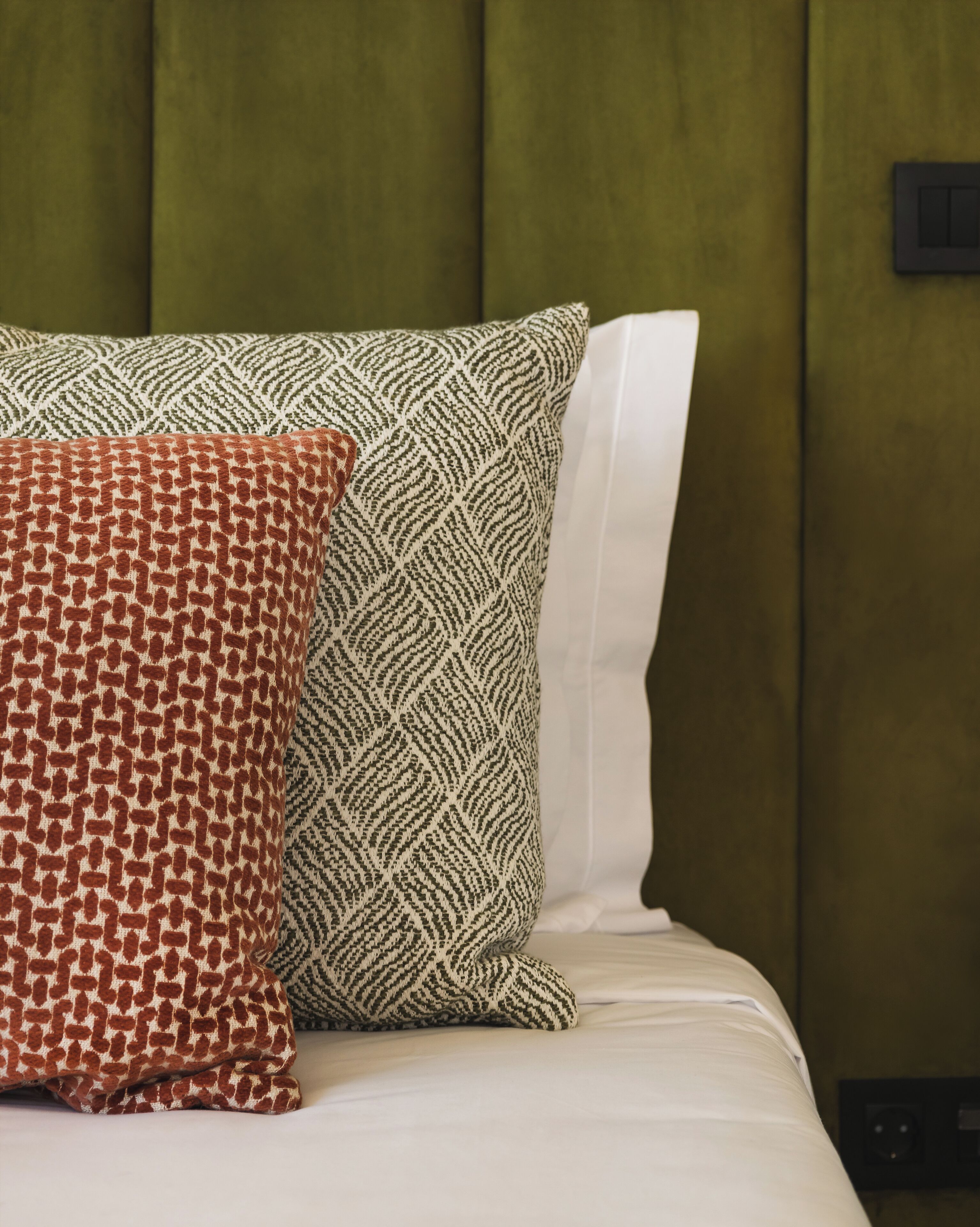 A bed with two pillows, one with a geometric pattern and the other with a zigzag pattern, against a green upholstered headboard.