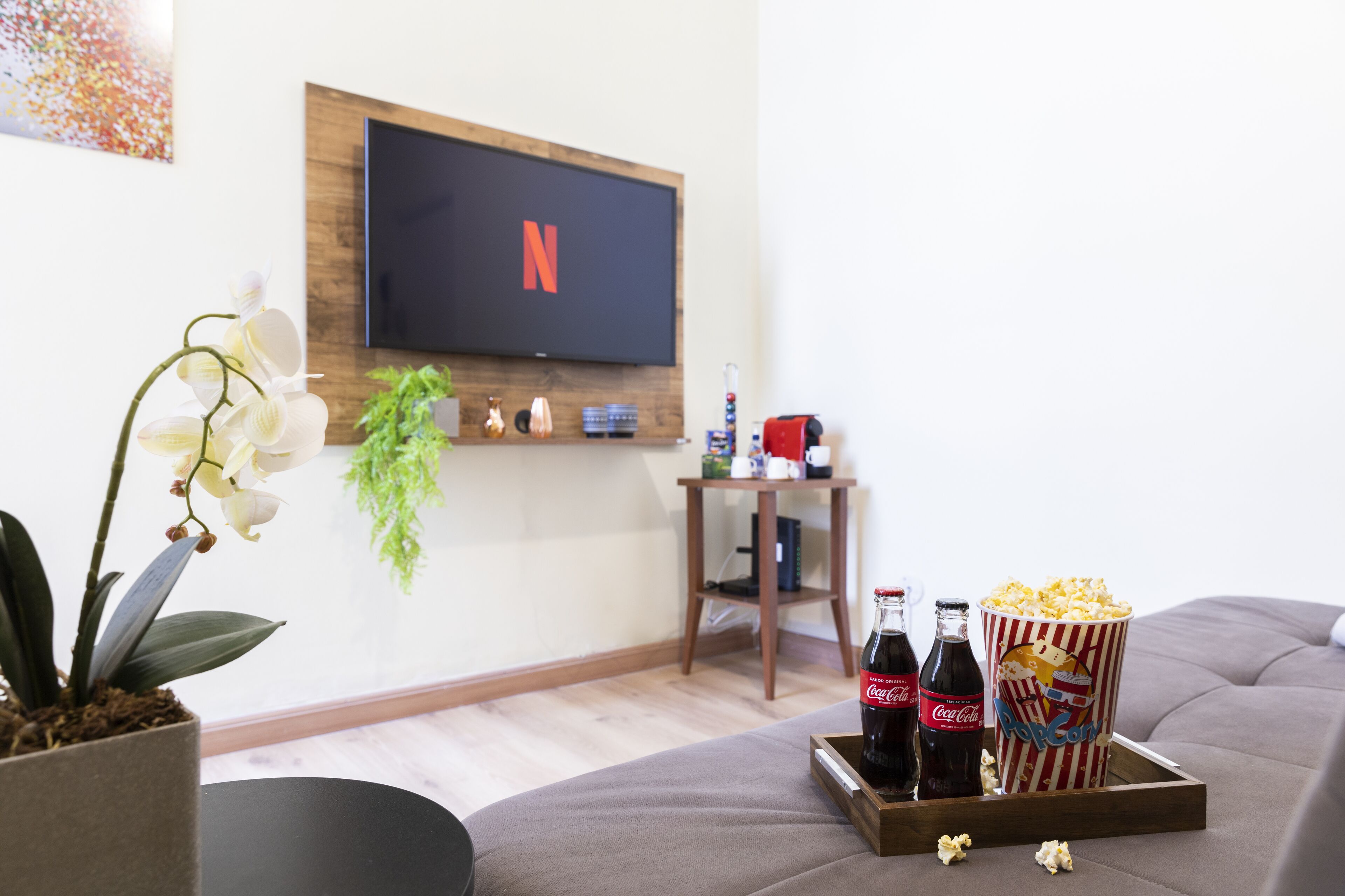 A living room with a flat-screen TV mounted on the wall, a wooden coffee table with popcorn and soda bottles, a gray sofa, and a potted orchid plant.