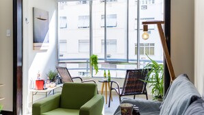 A modern living room with a green sofa, a wooden chair, and a small table with bottles. A large window offers a view of buildings outside. - Copacabana  (Rio de Janeiro)