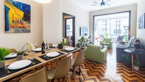 A dining area with a long table set for six, surrounded by chairs, with a painting on the wall, and a view of the outside through large windows. - Copacabana  (Rio de Janeiro)