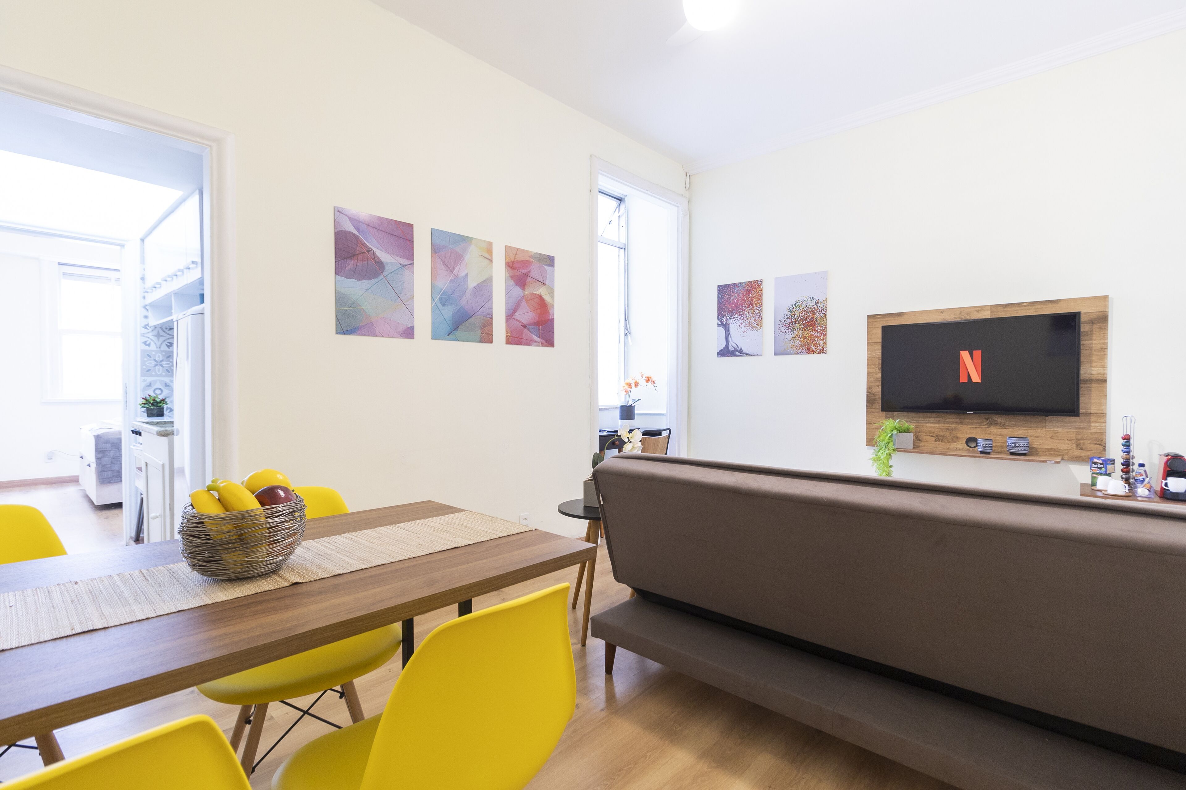 A modern living room with a wooden dining table, yellow chairs, a brown sofa, and a flat-screen TV mounted on the wall.