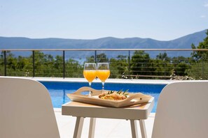 Ein Tisch am Pool mit zwei Orangensäften und einem Teller Croissants, mit Blick auf einen Pool und eine malerische Berglandschaft.