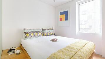Una cama bien hecha con almohadones estampados, una obra de arte abstracta enmarcada en la pared y una ventana con vistas a edificios.