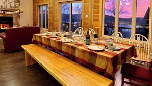 A long wooden dining table set for six with a bench, large windows, and a stone fireplace. - L'expérience Arborum Le Sime, Chalet en Bois Massif à L'emplacement Unique! (Saint-Léonard-de-Portneuf)