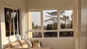 Une pièce d’angle avec un canapé d’angle beige, de grandes fenêtres donnant sur des arbres et des immeubles, et un plancher en bois.