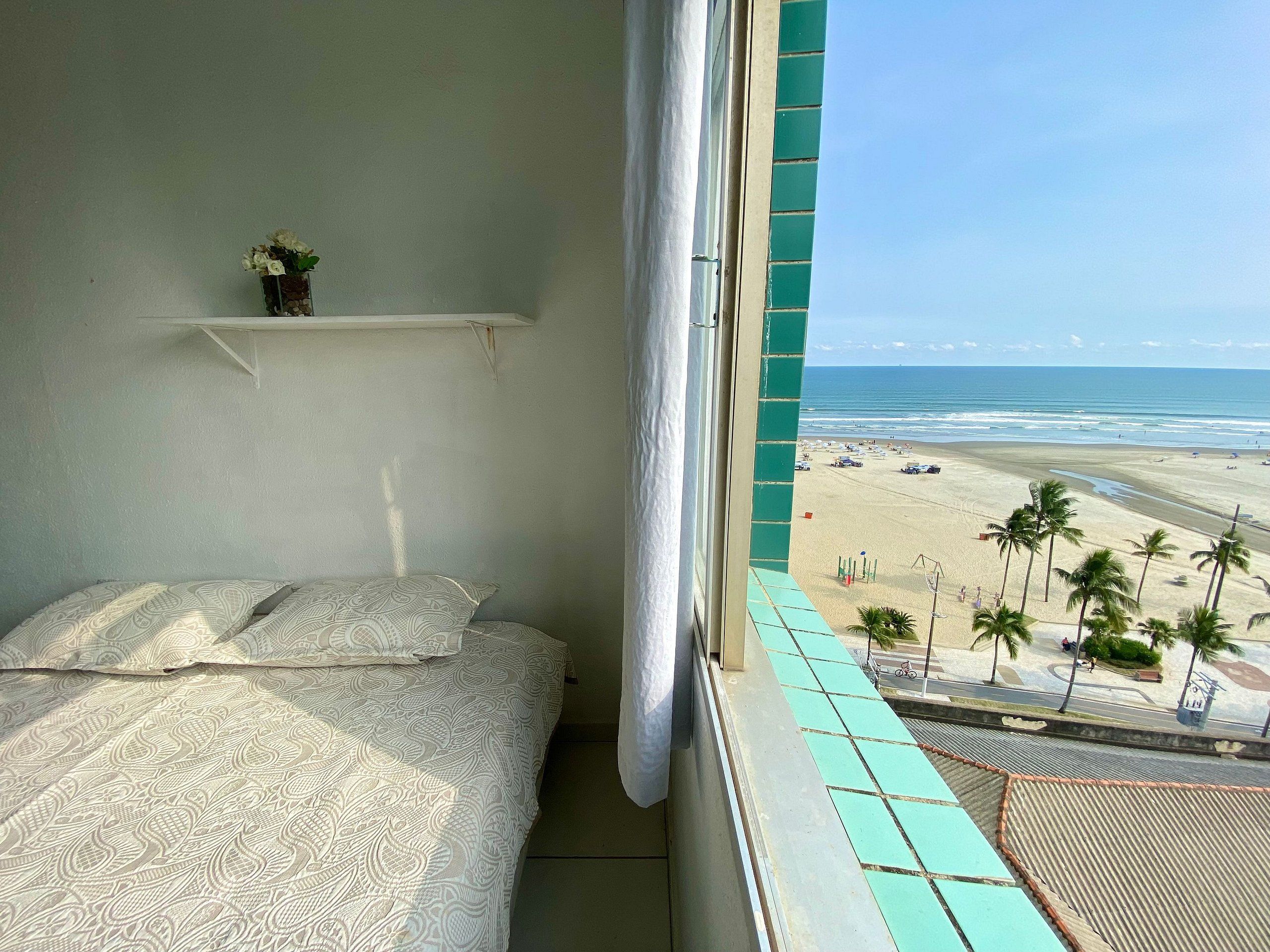 A bedroom with a bed, a small shelf with a vase, and a view of a beach with palm trees.