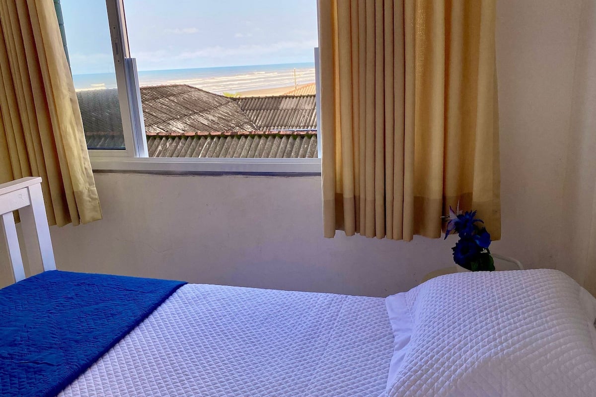A room with a bed, a window with curtains, a view of the beach, and a small plant.