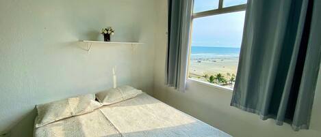 A bedroom with a bed, a window with curtains, a shelf with a vase, and a view of the beach.