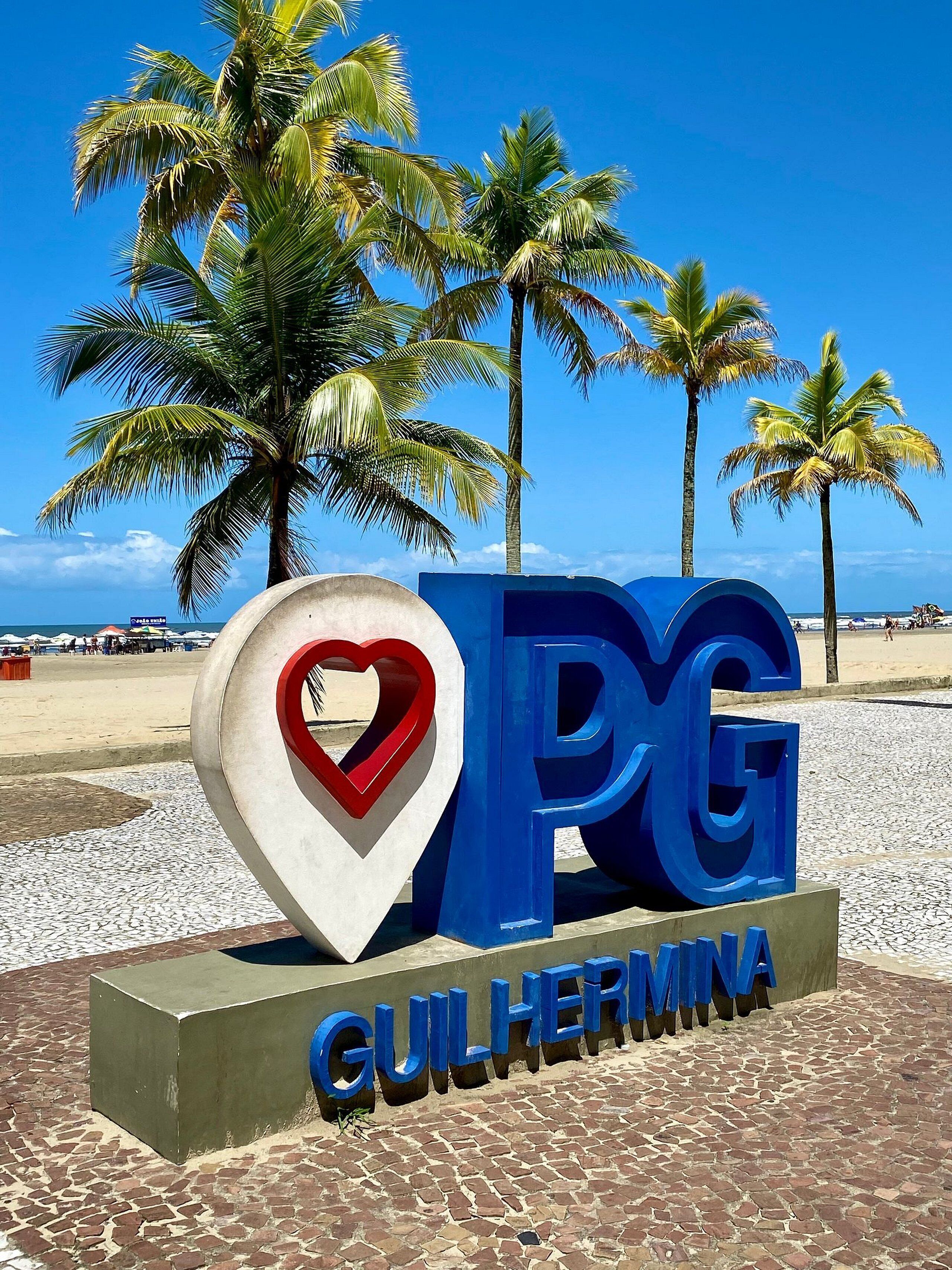 Un letrero con 'PG' y 'GUILHER' que indica una ubicación, con palmeras y una playa al fondo.