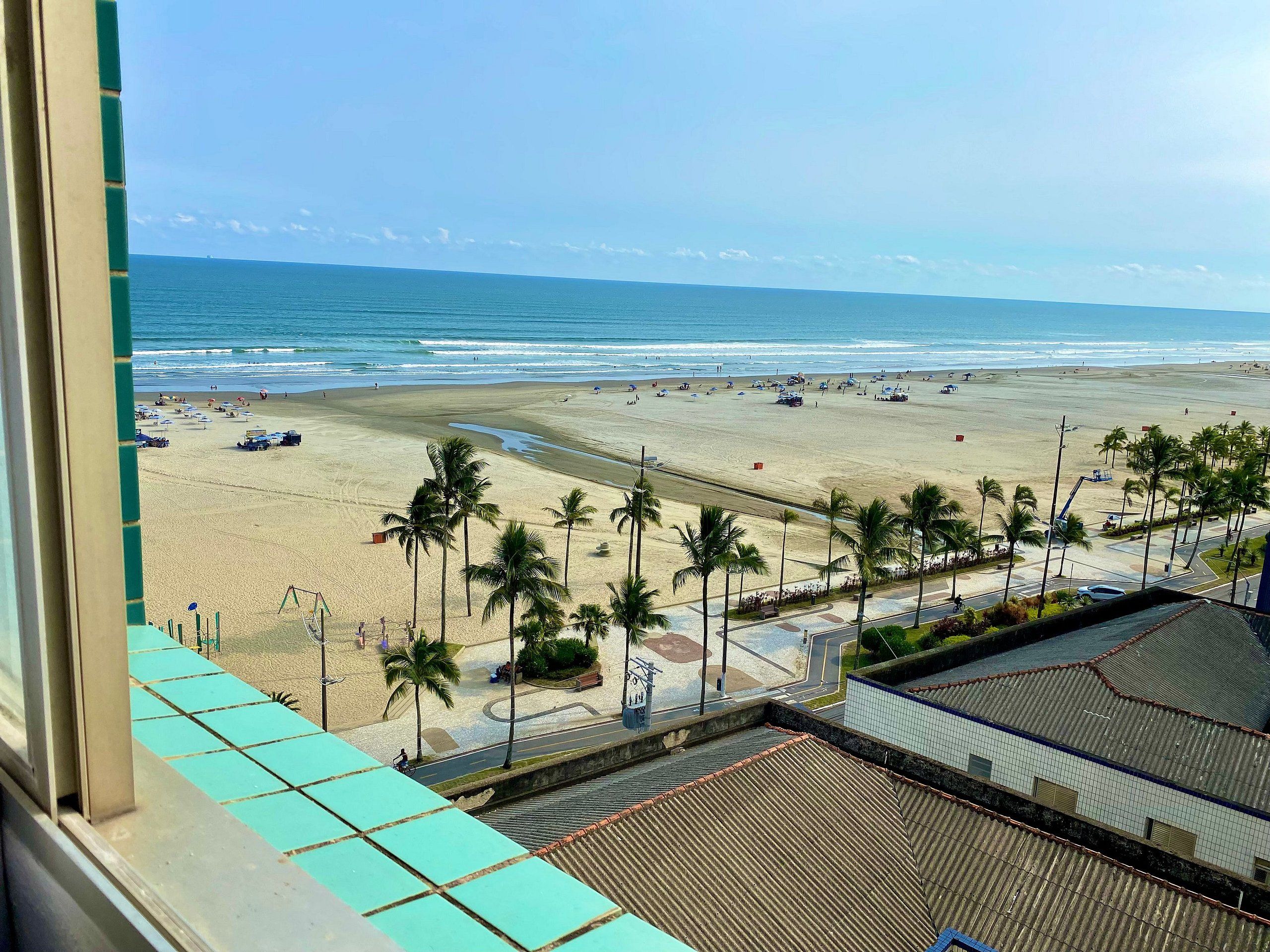 A beach view from a window overlooking a sandy beach with palm trees, a road, and a building with a tiled roof.