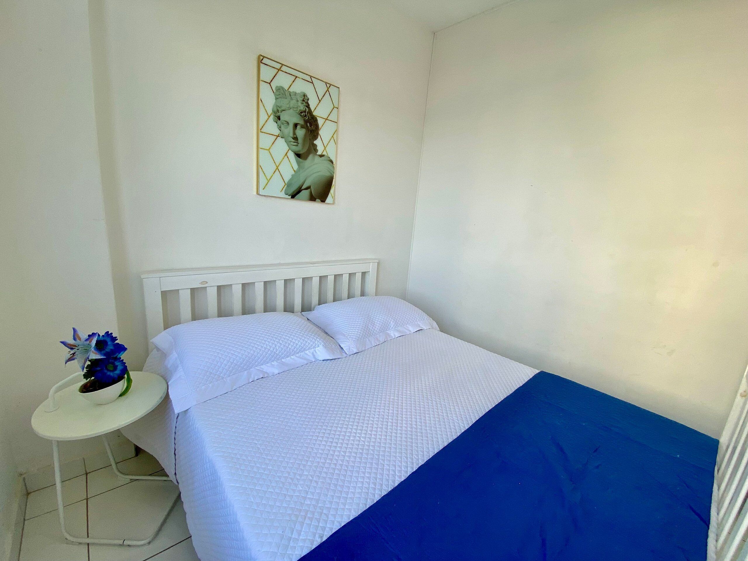 Quarto com cama, uma mesinha de cabeceira, um vaso com flores e um quadro na parede.