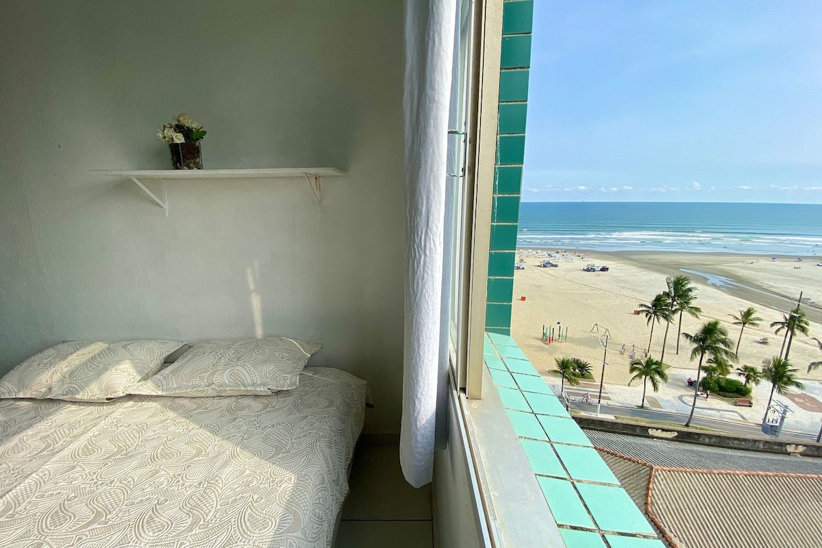 A bedroom with a bed, a small shelf with a vase, and a view of a beach with palm trees.