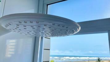 Eine an der Decke befestigte Duschbrause mit kreisförmigem Design, ein Fenster mit Blick auf den Strand und eine geflieste Wand.