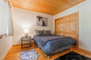 Ein Schlafzimmer mit Holzbalkendecke, einem Bett mit grauer Decke, einem kleinen Nachttisch mit Lampe, einem Fellteppich und einem Holzschrank.