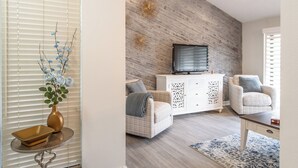 A living room with a modern design, featuring a wooden accent wall, a flat-screen TV, two armchairs, a white cabinet, and a small table with a vase and bowl.