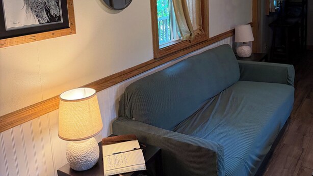 Una habitación con un sofá, una lámpara, un cuadro enmarcado de un pájaro, un espejo y una ventana con cortinas.