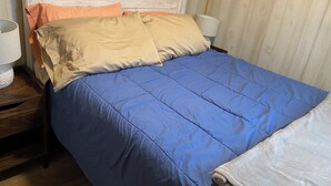 Una cama con colcha azul y dos almohadas color beige, cabecera de madera y mesita de noche con lámpara.