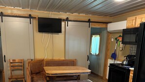Una habitación con un banco de madera, una mesa, un televisor montado en la pared y una zona de cocina con refrigerador y estufa.
