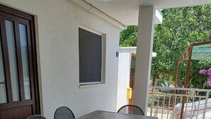 A covered outdoor dining area with a table and chairs, a window, and a view of greenery. - Studio Apartment Nikola (Janjina)