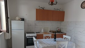 A compact kitchen with a refrigerator, stove, oven, and wooden cabinets. A dining table with chairs is set up in the center. - Studio Apartment Nikola (Janjina)