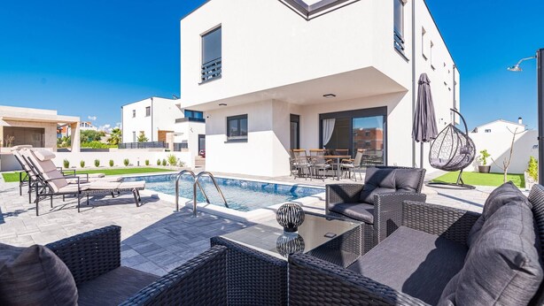 Un espace piscine extérieur moderne, doté de chaises longues, d’une table à manger et d’un fauteuil suspendu.