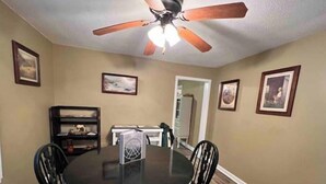 Un coin repas avec une table ronde, quatre chaises, un ventilateur de plafond et des tableaux encadrés accrochés aux murs.