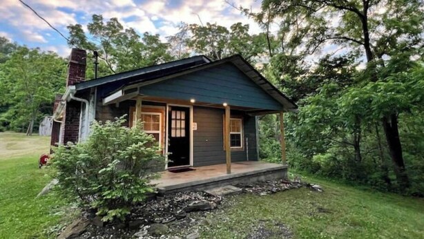 Une petite cabane à un seul étage, dotée d’un porche, entourée d’arbres et bordée d’une pelouse.