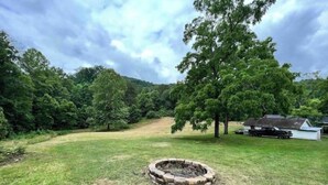 A grassy outdoor area with a circular stone fire pit, surrounded by trees and a small white building. - Cozy Country Home for all ATV Riders! Direct Access to Tackett and Royal Blue! (LaFollette)