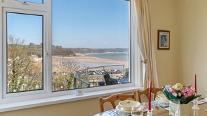 A dining area with a table set for two, a view of the beach, and a window with curtains. - White Sails - Beach and Harbour Views (Saundersfoot)