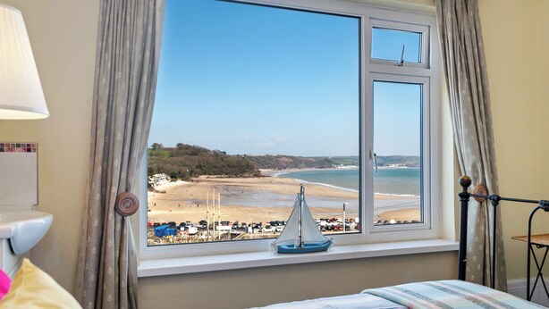 A room with a large window overlooking a beach, a bed with a striped bedspread, and a small sailboat decoration on the windowsill. - White Sails - Beach and Harbour Views (Saundersfoot)