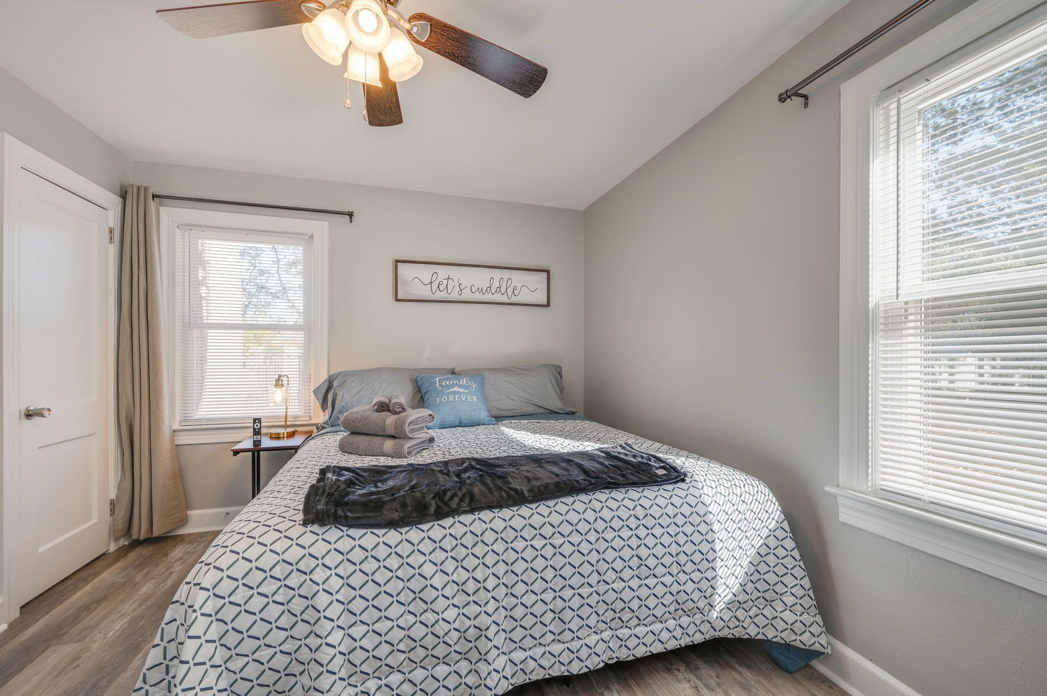 Une chambre à coucher comprenant un lit, un ventilateur de plafond, une fenêtre avec des stores, une table de chevet avec une lampe et un mur orné d’un tableau.