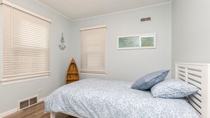 Ein Schlafzimmer mit einem Bett, zwei Fenstern, einer kleinen Holzablage und einem an der Wand hängenden Bild.