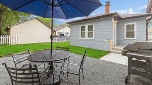 Eine Terrasse mit rundem Tisch und Stühlen, einem blauen Sonnenschirm und einem Grill.