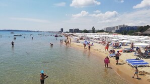 A beach with clear shallow waters, people swimming and sunbathing, and buildings in the background. - Billy's 2 bed apartment for Rent at Sun Village Complex (Слънчев бряг)