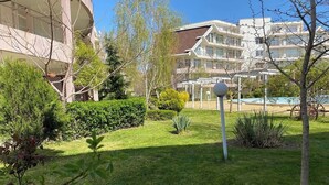 A well-maintained garden with a lawn, various shrubs, and a few trees. In the background, there are multi-story buildings with balconies. - Billy's 2 bed apartment for Rent at Sun Village Complex (Слънчев бряг)