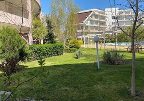 Ein gepflegter Garten mit Rasen, verschiedenen Sträuchern und einigen Bäumen. Im Hintergrund sind mehrstöckige Gebäude mit Balkonen zu sehen.