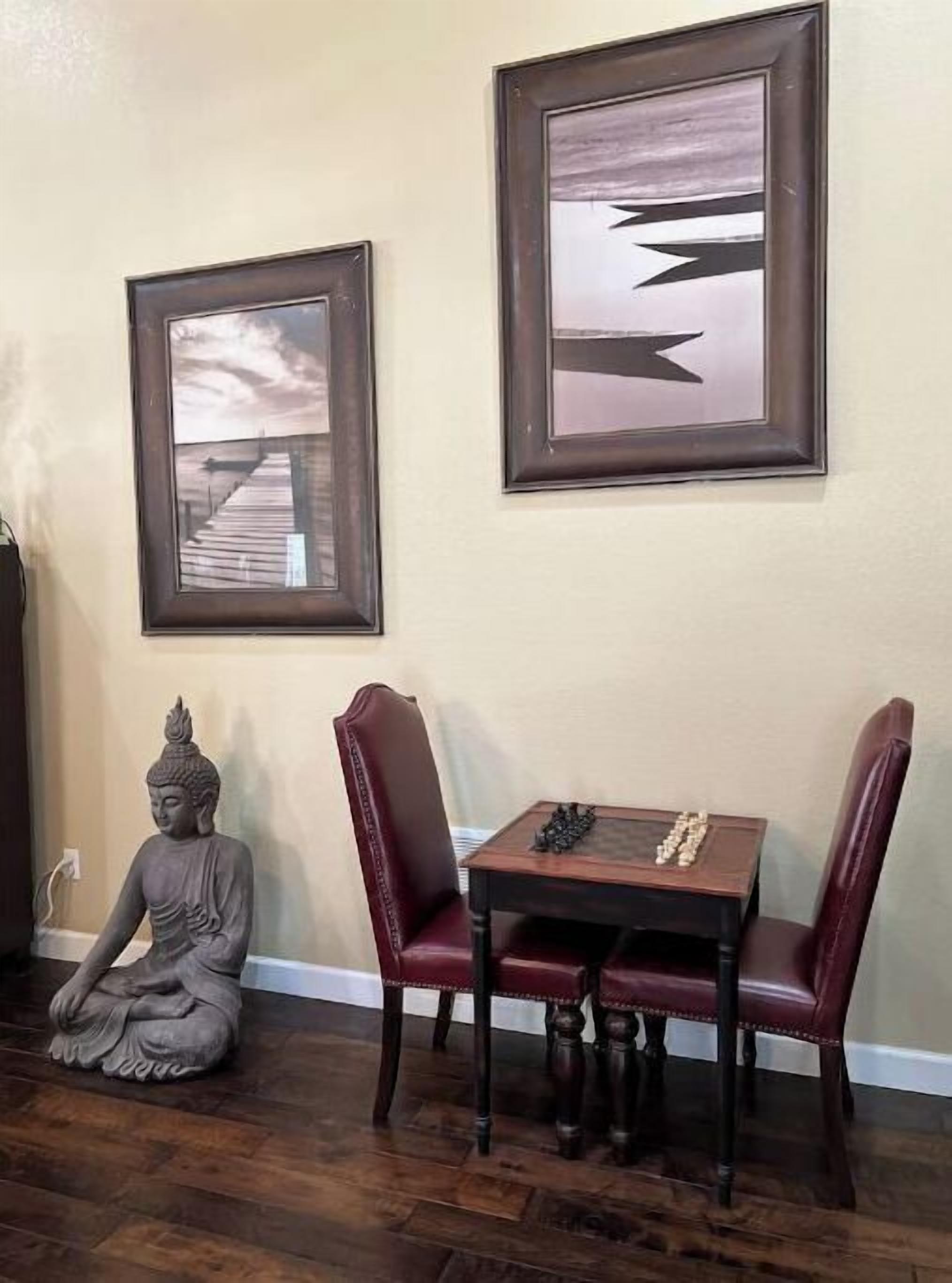 Um quarto com piso de madeira, uma estátua de Buda, uma mesa com peças de xadrez e duas pinturas abstratas emolduradas na parede.